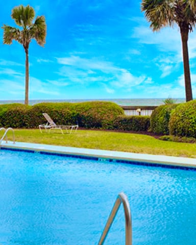 Swimming pool with lounge chairs, palm trees, and manicured hedges under blue sky