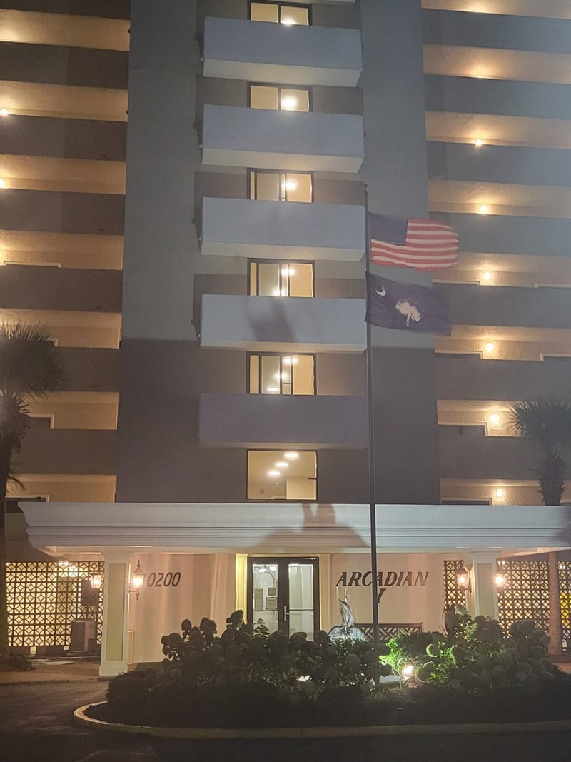 Modern multi-story hotel building at night with illuminated balconies, ground-floor entrance, and American flag displayed on facade