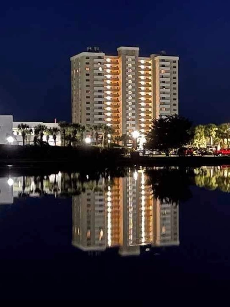 Illuminated high-rise residential building reflected in calm water at night with palm trees and dark blue sky