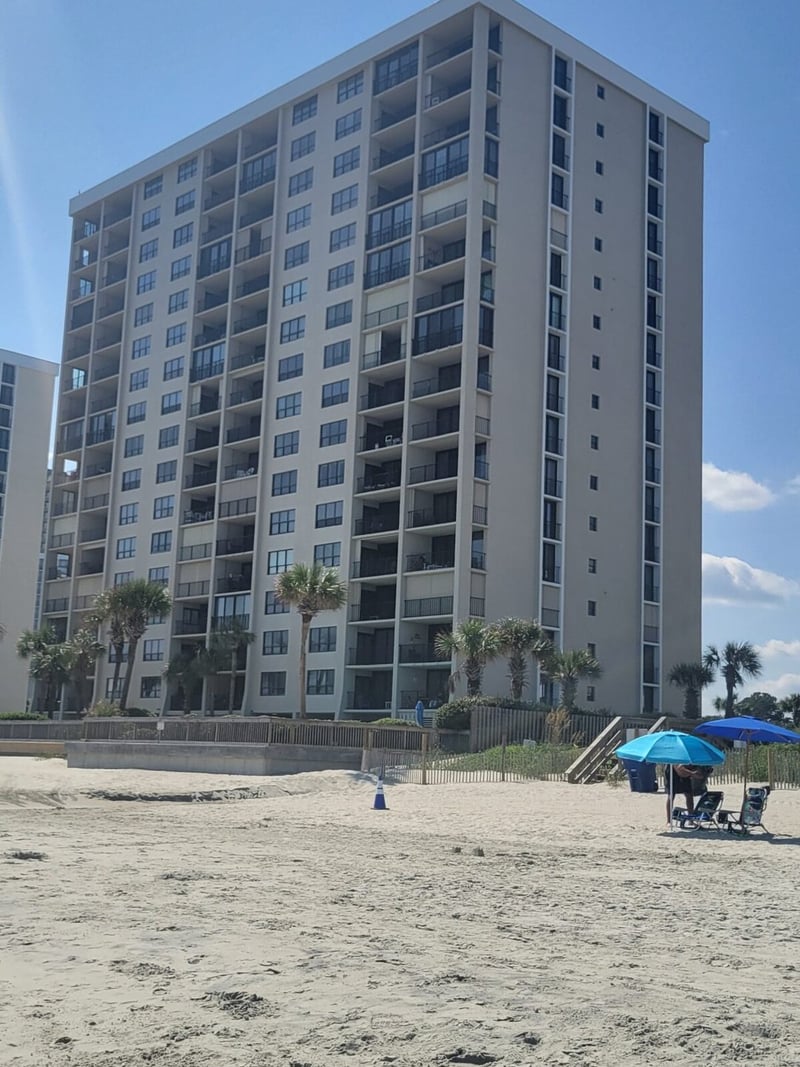 Multi-story beachfront condominium building with gray facade and balconies, viewed from sandy beach with palm trees and blue umbrella in foreground