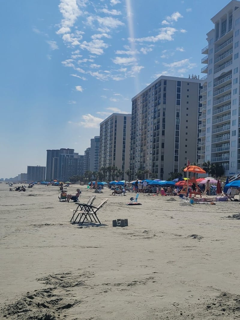 Sandy beach with sunbathers and umbrellas, high-rise residential buildings lining the shore, blue sky with white clouds