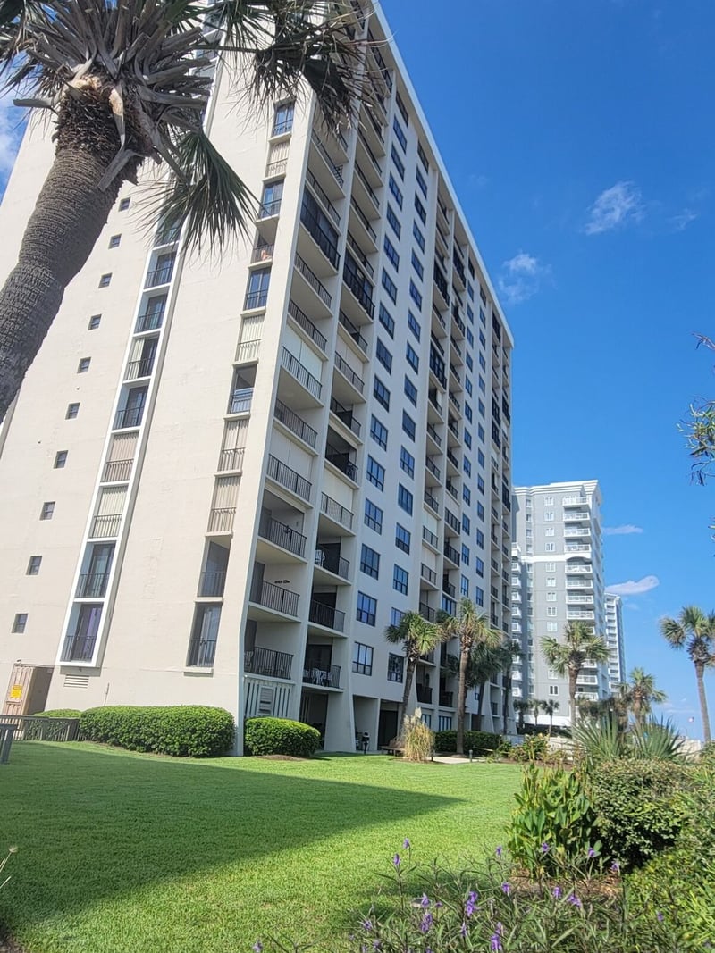 Multi-story white residential building with palm trees, green lawn, and purple flowering plants in foreground under blue sky