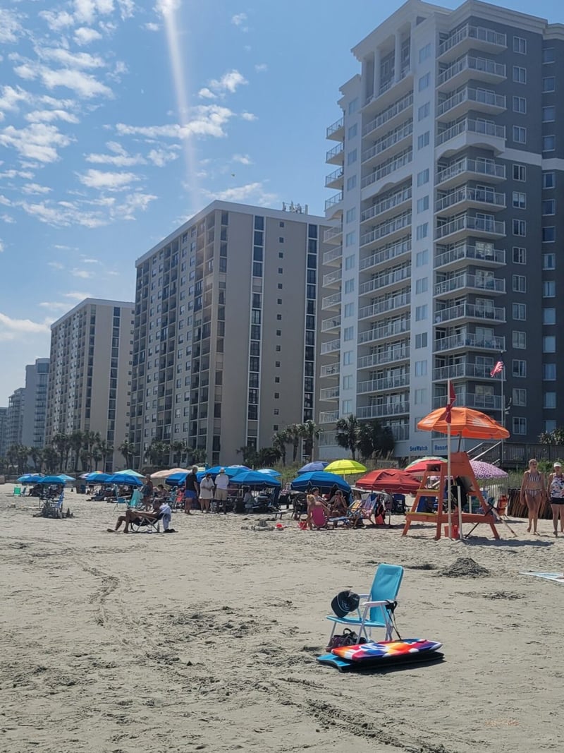 Beach with colorful umbrellas and beachgoers in front of tall residential buildings under bright sunny sky