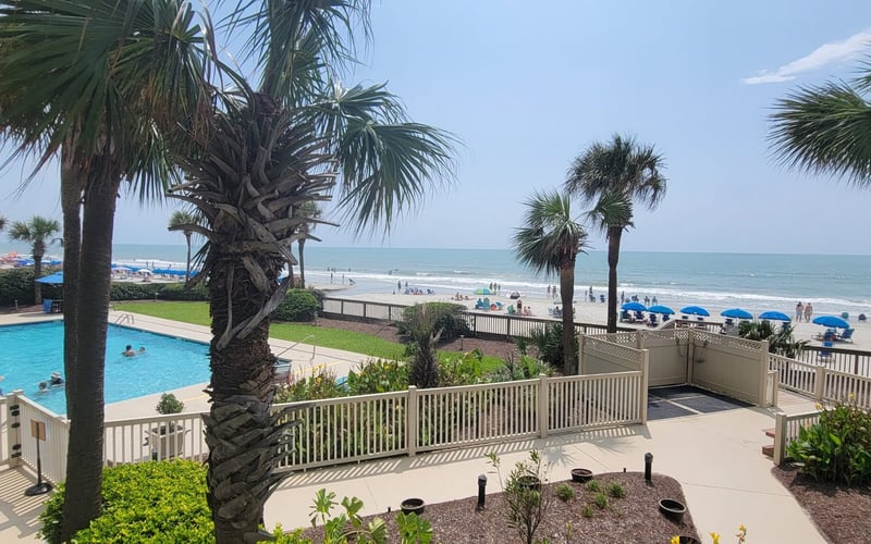 Beachfront resort with swimming pool, palm trees, and ocean view with blue umbrellas on sandy beach