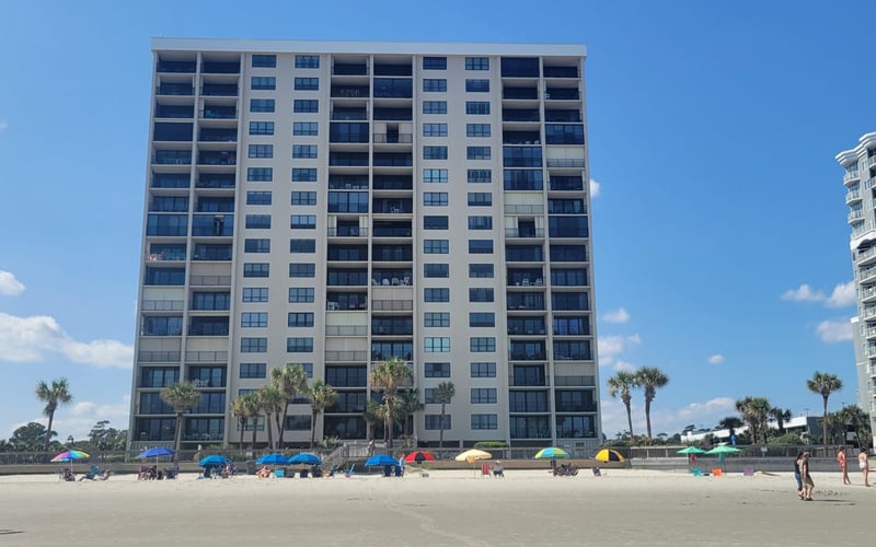 Beachfront high-rise condominium building with sandy beach, colorful umbrellas, and palm trees
