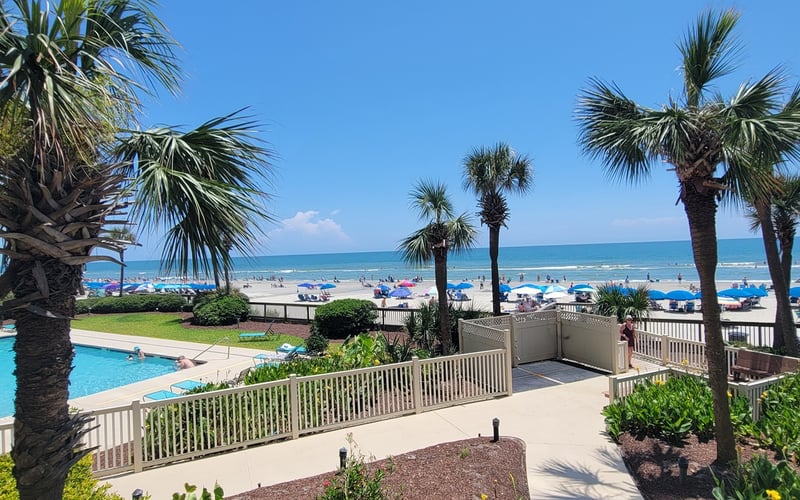 Beachfront resort with swimming pool, palm trees, white railings, and ocean view with colorful umbrellas on sand