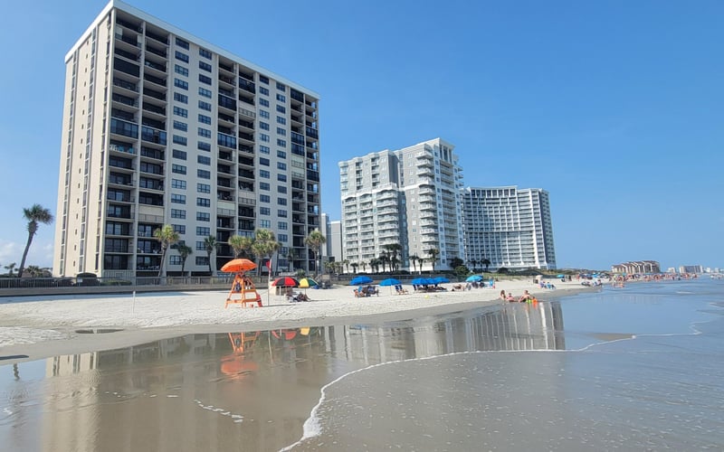 Beachfront high-rise condominiums with sandy beach, shallow water, and beachgoers under umbrellas
