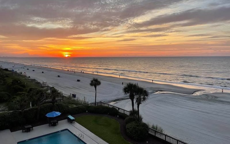 Oceanfront property at sunset with swimming pool, palm trees, and sandy beach with scattered beachgoers