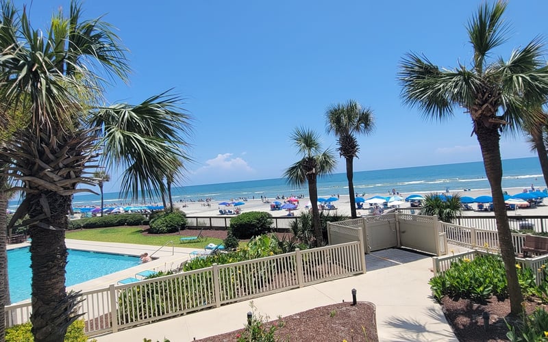 Beachfront resort with swimming pool, palm trees, and ocean view with blue umbrellas on sandy beach