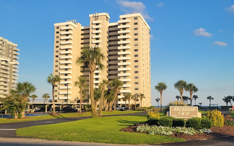 Beachfront condominium complex with multiple tan high-rise towers, palm trees, and landscaped grounds under clear blue sky