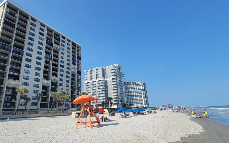 Beachfront with high-rise condominiums, lifeguard stand with orange umbrella, and swimmers in ocean water