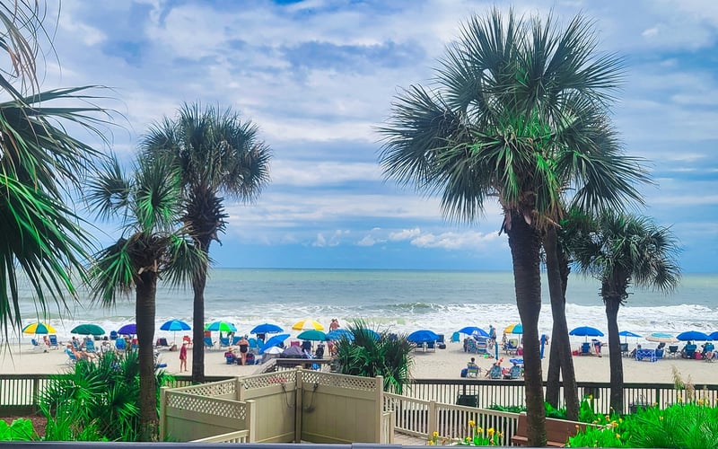 Beach view with palm trees, colorful umbrellas, and beachgoers from elevated deck with railing