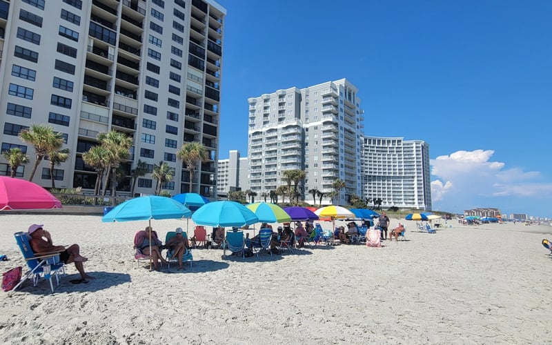 Sandy beach with colorful umbrellas and beachgoers, high-rise condominiums and palm trees in background