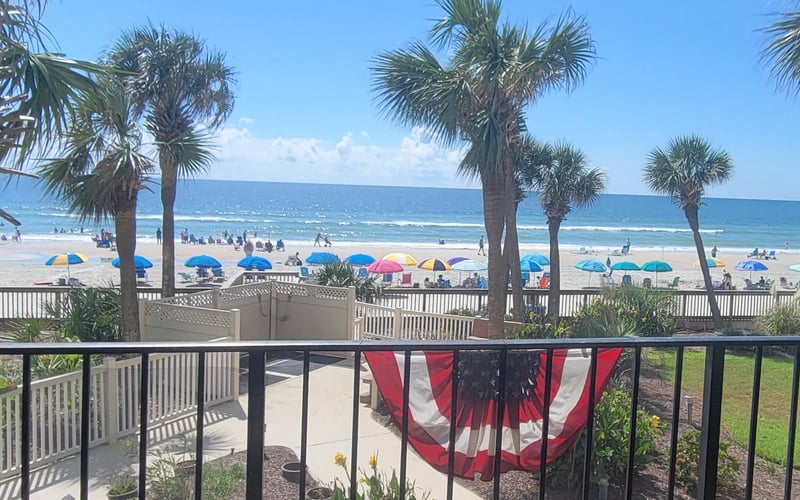 Beach view from oceanfront deck with palm trees, colorful umbrellas, and swimmers in the water