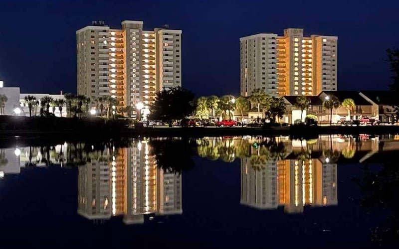Illuminated waterfront apartment towers reflected in calm water at dusk with palm trees and low buildings in foreground